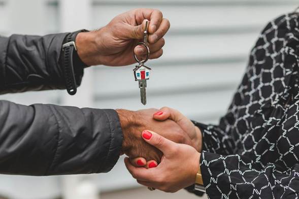 A person hands over a house key with a house-shaped keychain while shaking hands with another person, symbolizing a real estate transaction or new home purchase. | RealEstateMarket