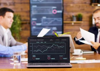 Four people sit at a table in a meeting room with a laptop displaying an annual report graph. Charts are visible on a large screen in the background. Papers and drinks are on the table. | RealEstateMarket
