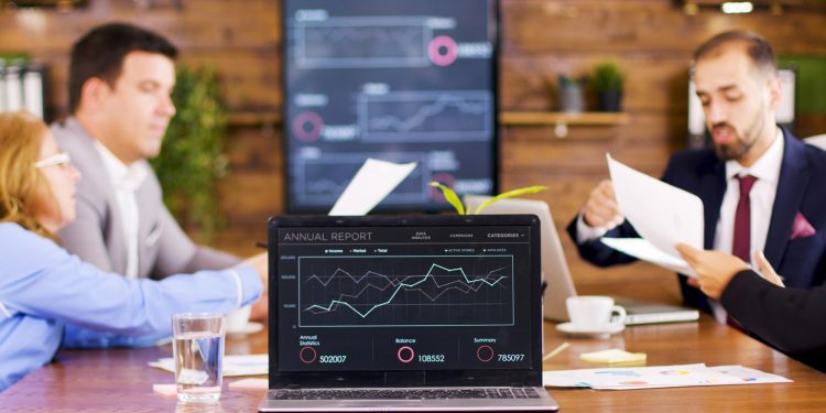 Four people sit at a table in a meeting room with a laptop displaying an annual report graph. Charts are visible on a large screen in the background. Papers and drinks are on the table. | RealEstateMarket