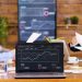 Four people sit at a table in a meeting room with a laptop displaying an annual report graph. Charts are visible on a large screen in the background. Papers and drinks are on the table. | RealEstateMarket