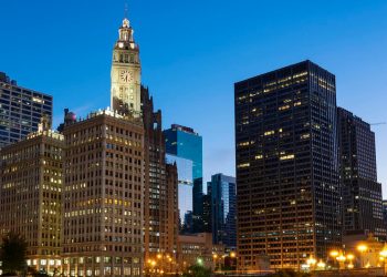 City skyline at dusk with illuminated office buildings and a prominent clock tower, under a clear blue sky. | RealEstateMarket