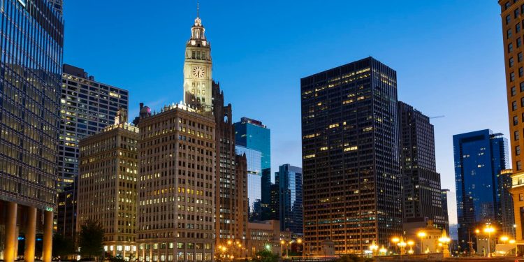 City skyline at dusk with illuminated office buildings and a prominent clock tower, under a clear blue sky. | RealEstateMarket