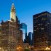 City skyline at dusk with illuminated office buildings and a prominent clock tower, under a clear blue sky. | RealEstateMarket