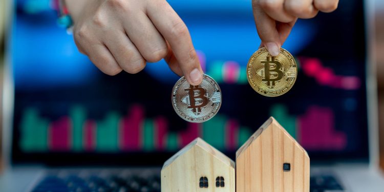 Hands holding a silver and a gold Bitcoin coin above two small wooden house models in front of a laptop screen displaying stock market charts. | RealEstateMarket