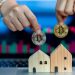 Hands holding a silver and a gold Bitcoin coin above two small wooden house models in front of a laptop screen displaying stock market charts. | RealEstateMarket
